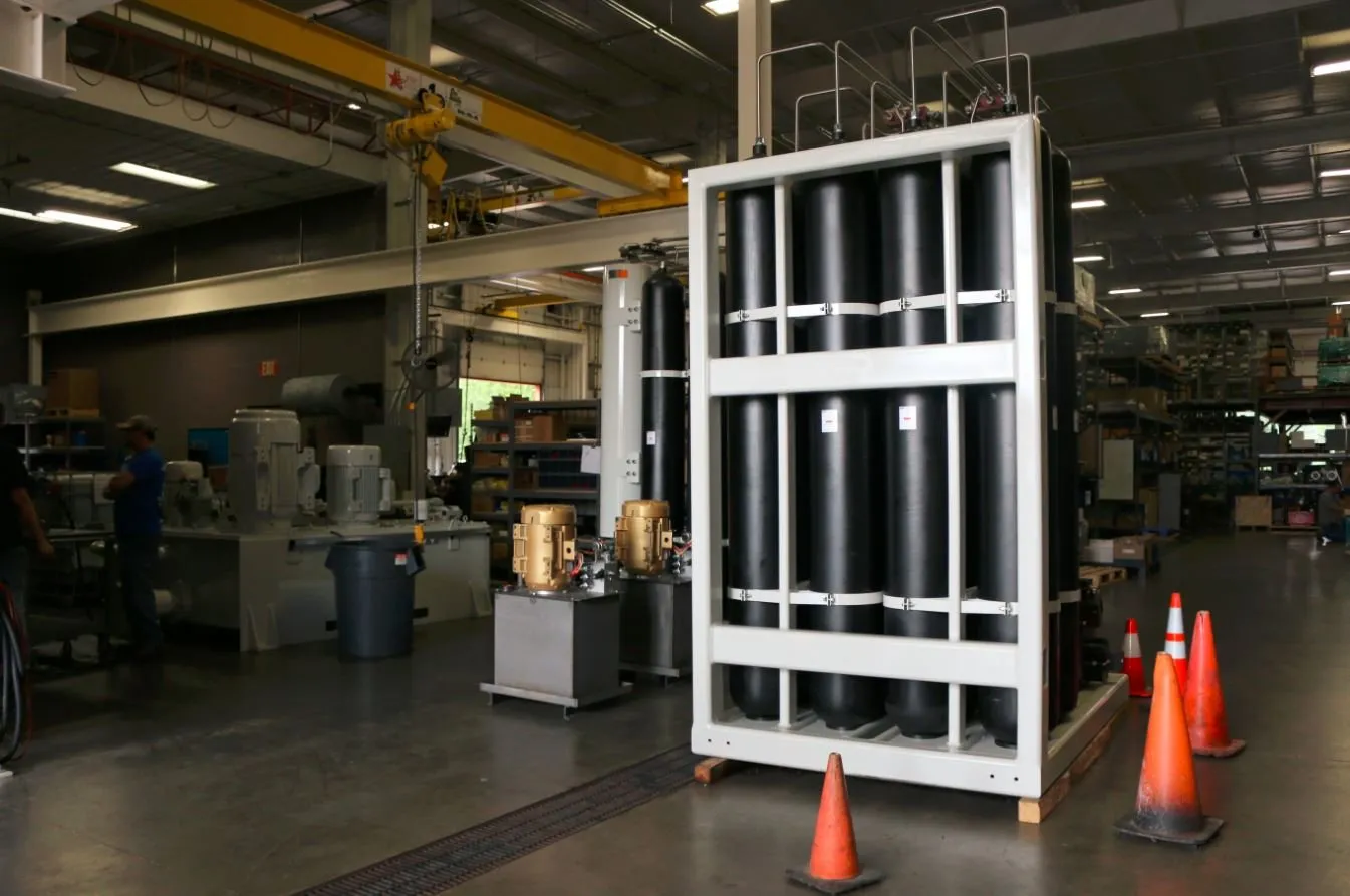 Rack of eight black cylindrical accumulators in a white steel frame, with safety cones and workers in the background