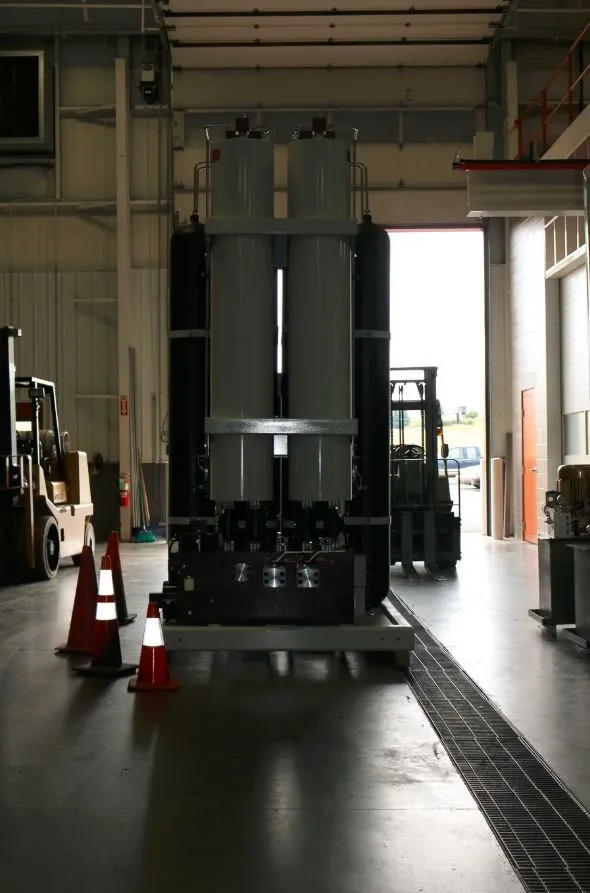 Two large cylindrical accumulators on a steel frame, viewed from across a warehouse floor with forklifts nearby