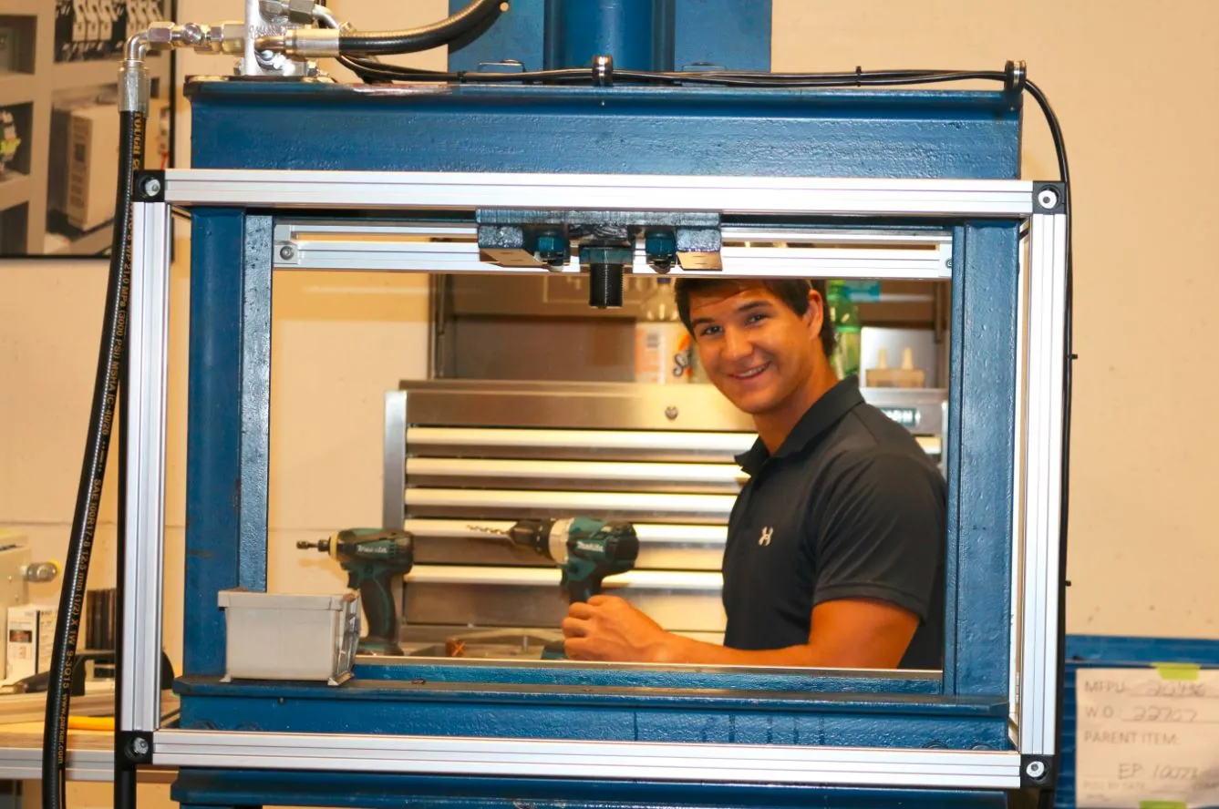 Man smiling through the window of a blue steel test fixture fitted with cordless drills and a hydraulic hose