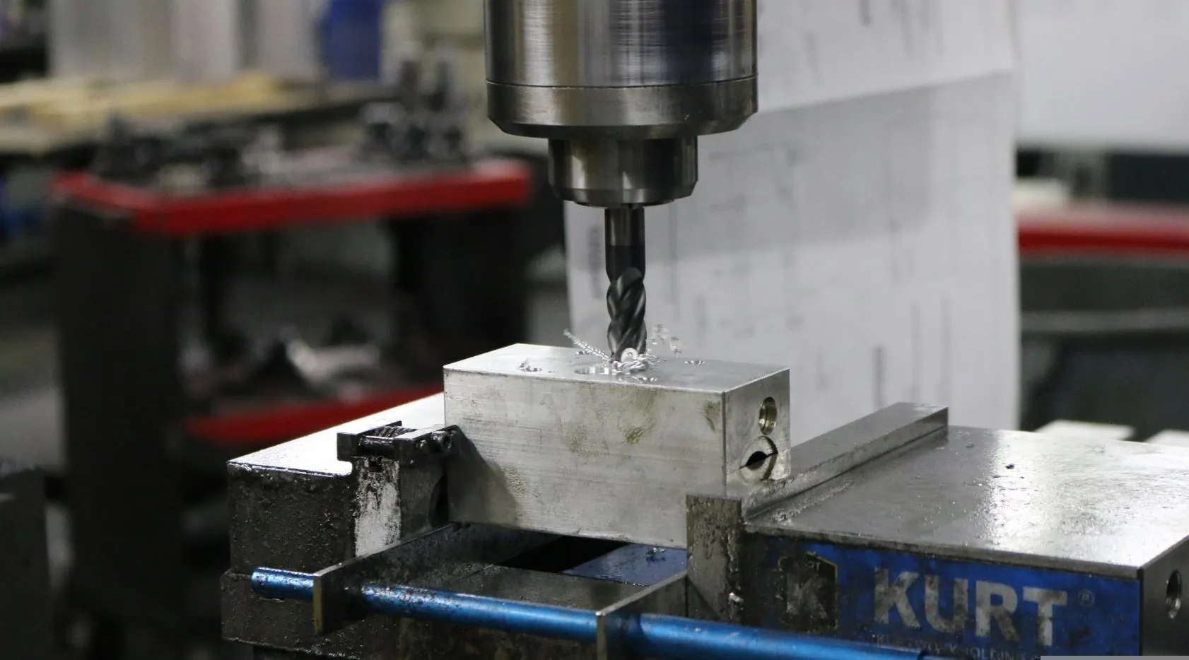 An end mill cuts an aluminum manifold block held in a blue Kurt vise