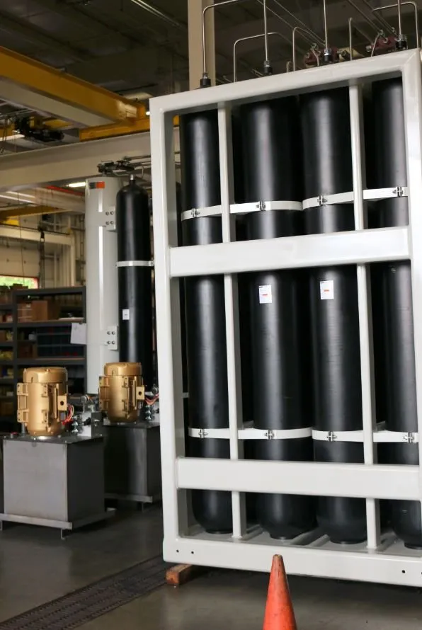 Rack of eight black cylindrical accumulators in a white steel frame, with a safety cone and two motors in the background