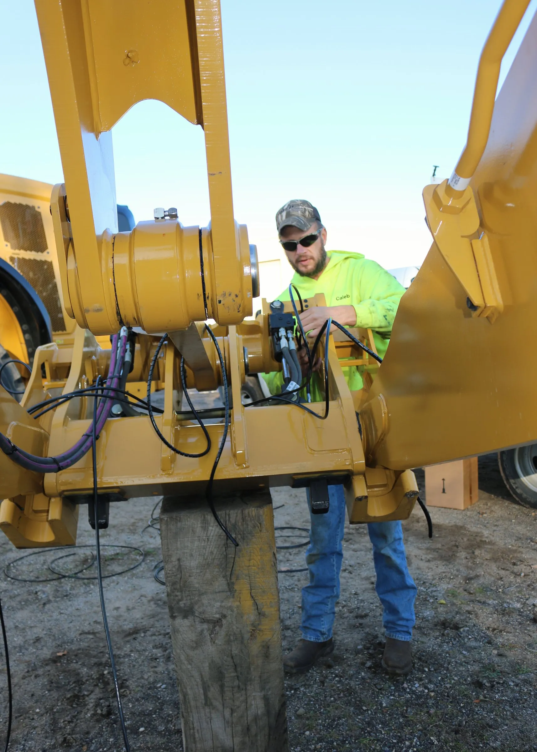 Man in a hi-vis jacket adjusting a hydraulic cylinder pin on a yellow excavator arm outdoors