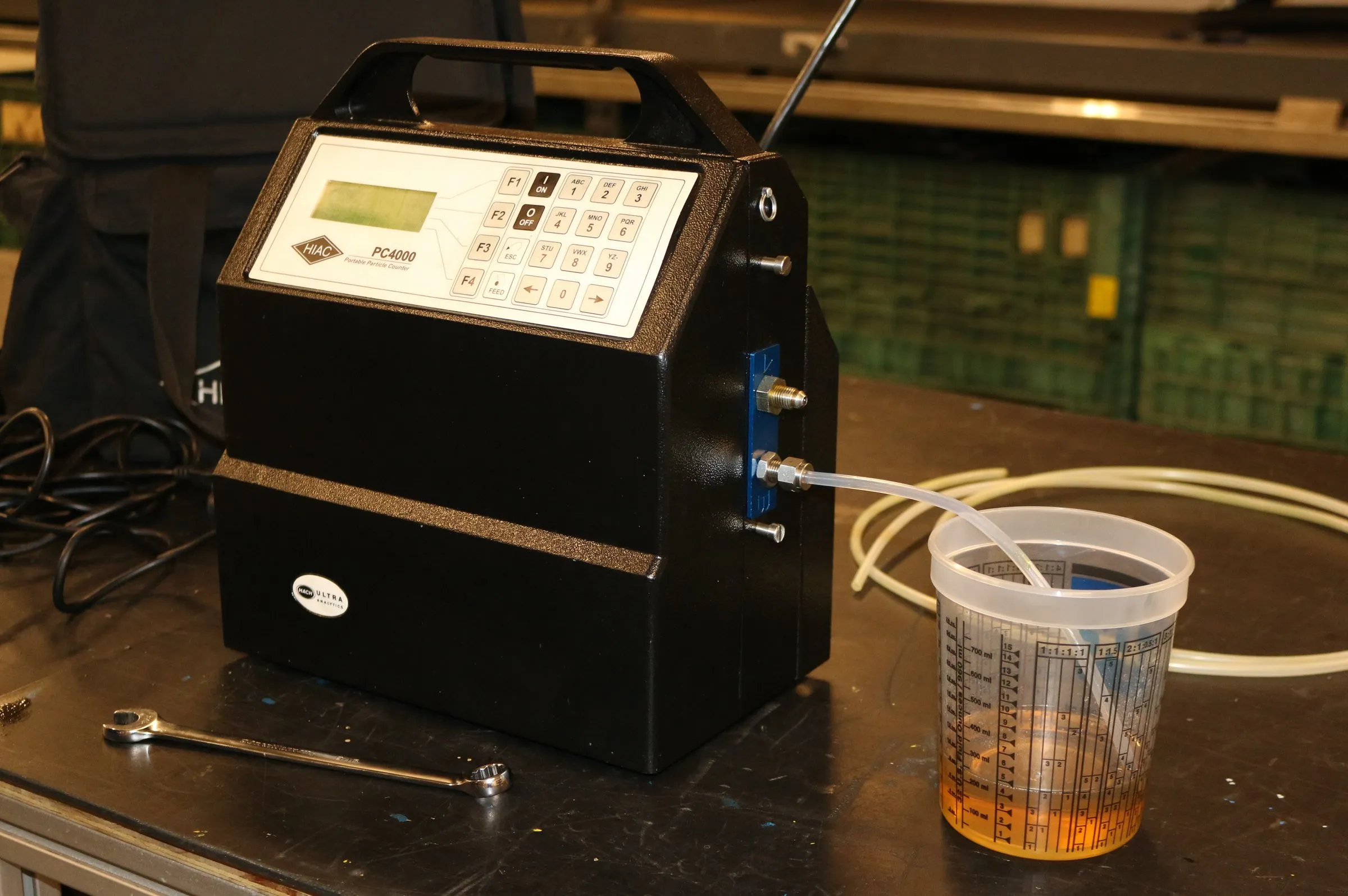 Portable particle counter connected by tube to a cup of amber hydraulic oil for sample testing