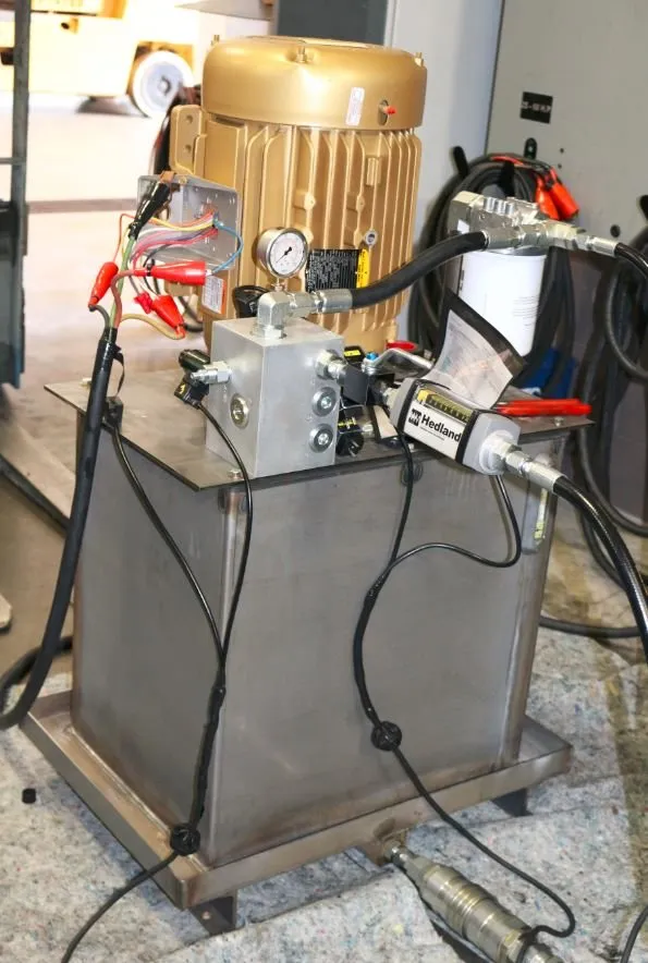 Gold-painted motor and Hedland flow meter on a stainless hydraulic reservoir, with a forklift blurred behind it