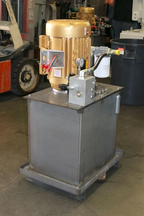 Gold-painted motor mounted on a hydraulic reservoir, with a forklift tire and a trash can nearby