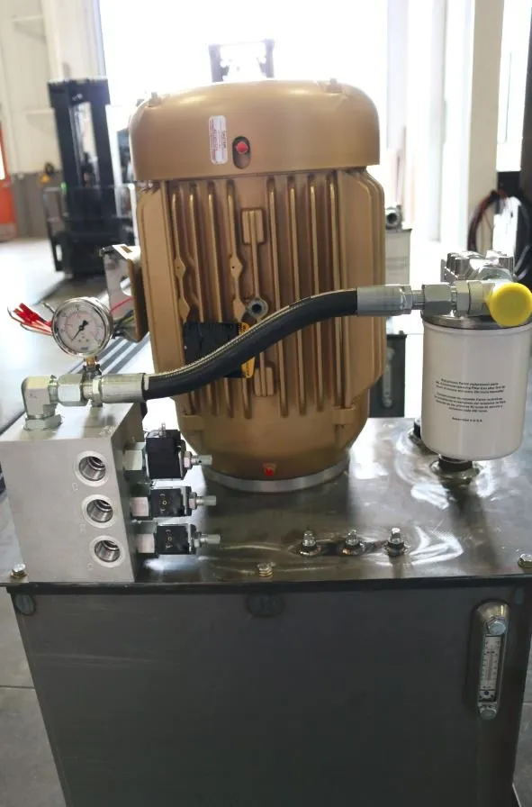Gold-painted motor mounted on a hydraulic reservoir with a pressure gauge and hose, in a shop with a forklift in the background