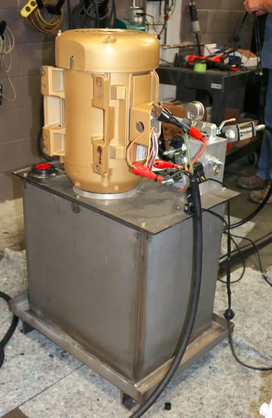 A gold electric motor on a stainless steel hydraulic reservoir, wired with alligator-clip test leads