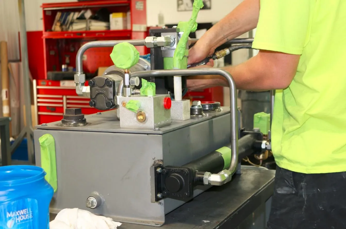 Hands fit a valve block onto a small hydraulic power unit on a workbench, marked with green tape flags
