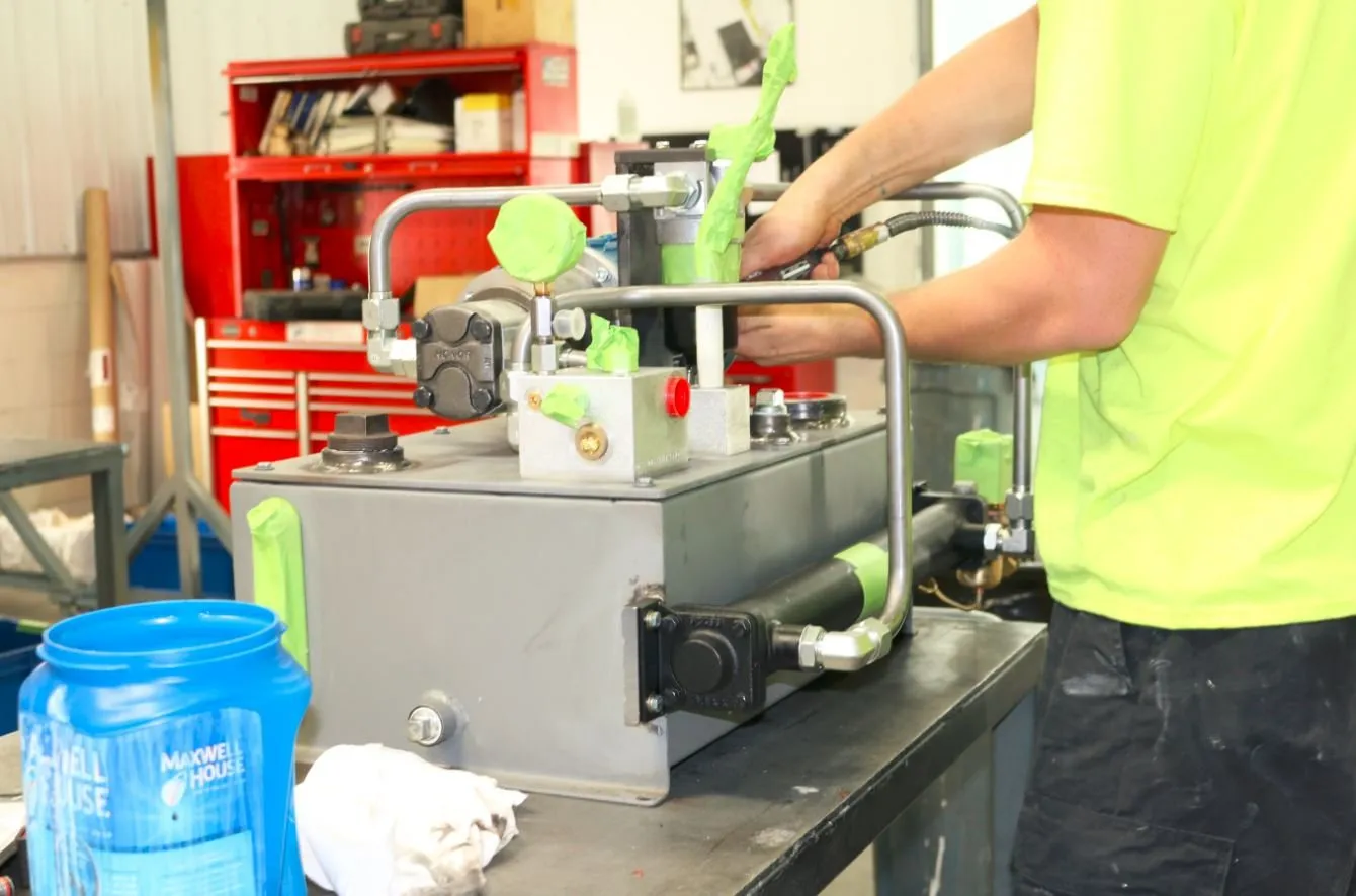Hands fit a valve block onto a small hydraulic power unit on a workbench, marked with green painter's tape flags