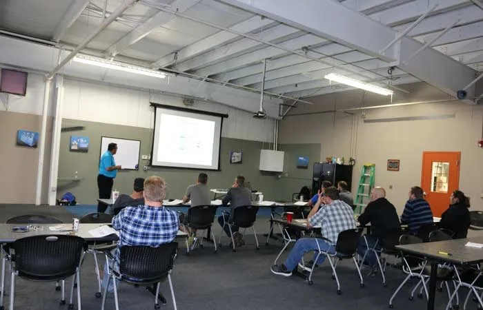 Instructor points to a projector screen while a class takes notes at tables