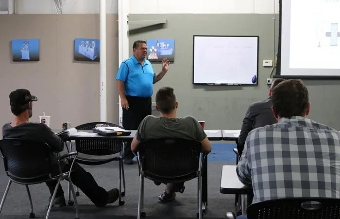 Instructor presents to a seated class in a training room with a whiteboard and projector