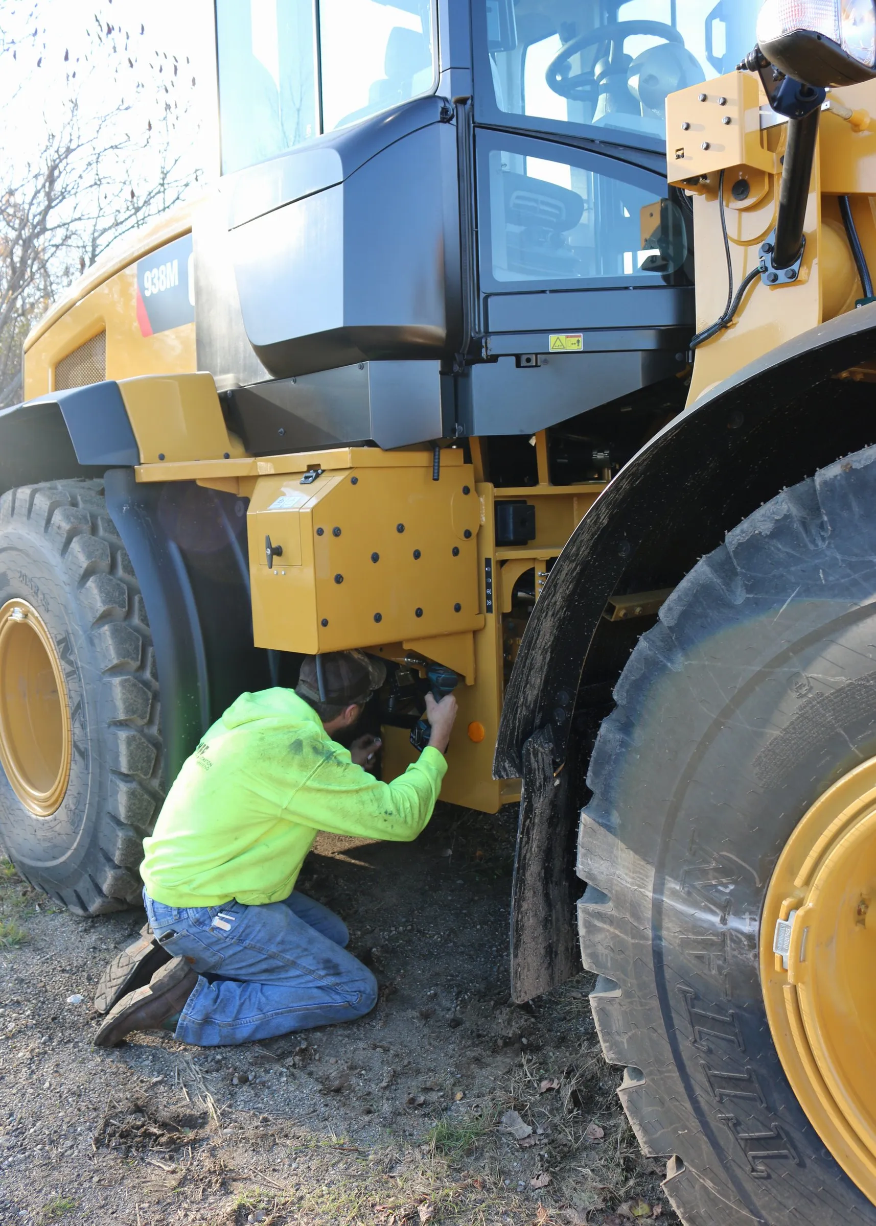 Technician kneeling under the front axle of a Caterpillar wheel loader making an adjustment