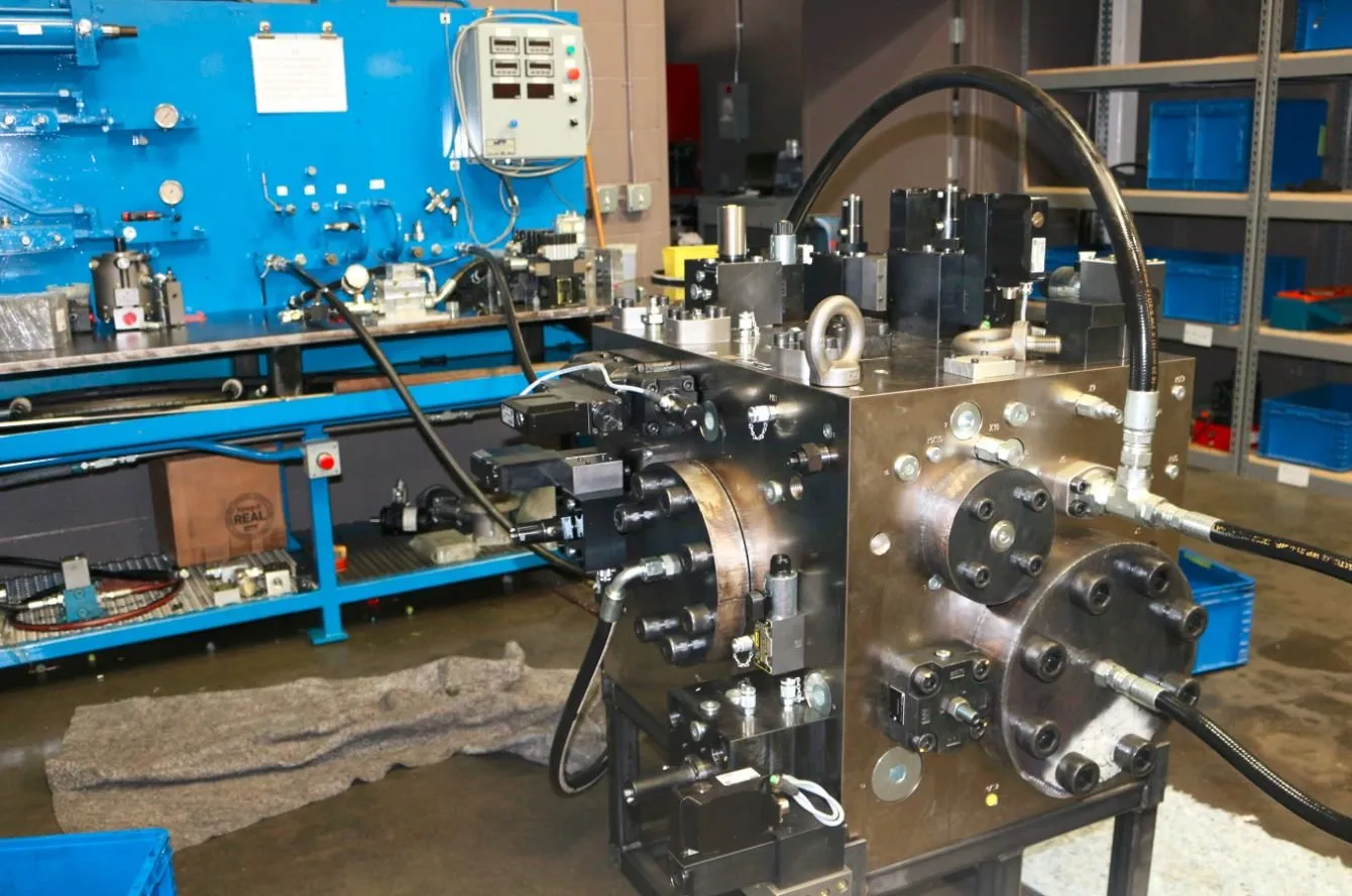 A large black hydraulic manifold block with multiple valves and gauges, set up on a test stand beside a blue test bench