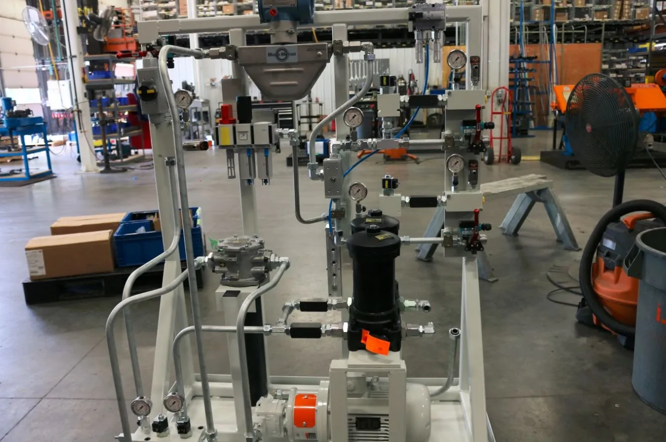 A hydraulic metering skid with valve manifolds and gauges, viewed from the rear near the motor and pump