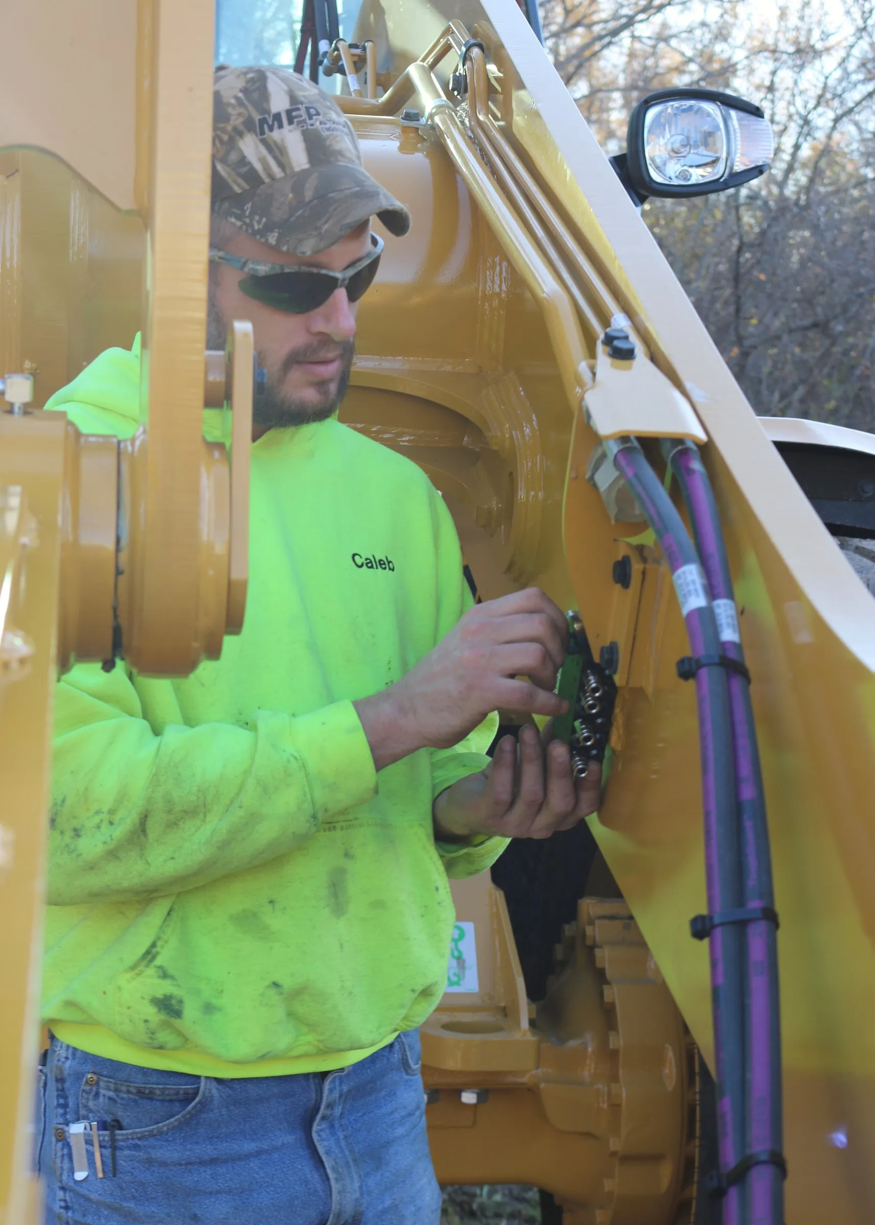 Technician in an MFP cap connects wiring near hydraulic lines on a wheel loader