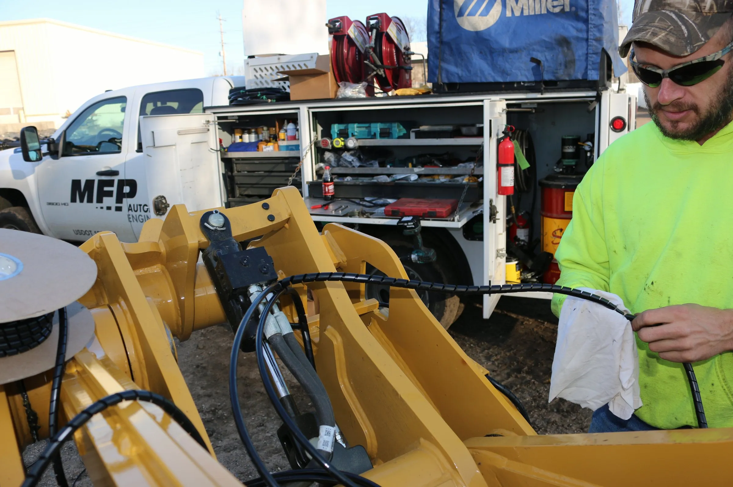 A worker wipes a hydraulic hose fitting on a yellow excavator attachment beside an MFP service truck outdoors