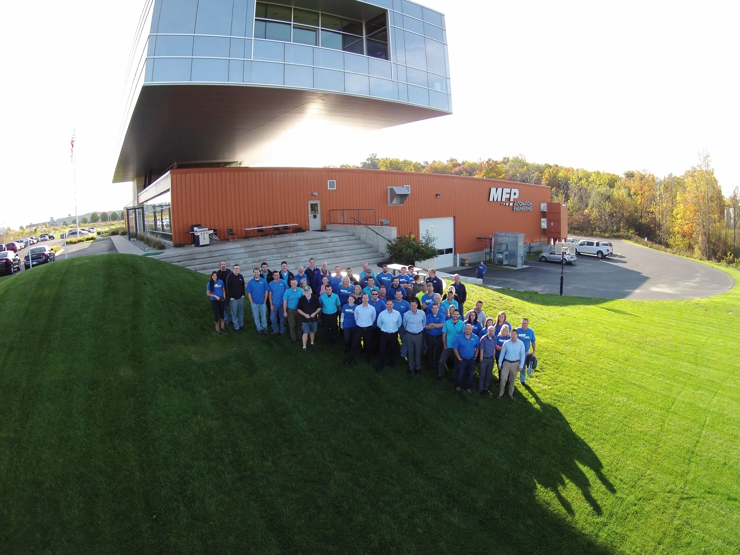 Aerial photo of a large group of people, several in MFP shirts, on the lawn in front of MFP's orange warehouse and glass office wing in Hudsonville, MI