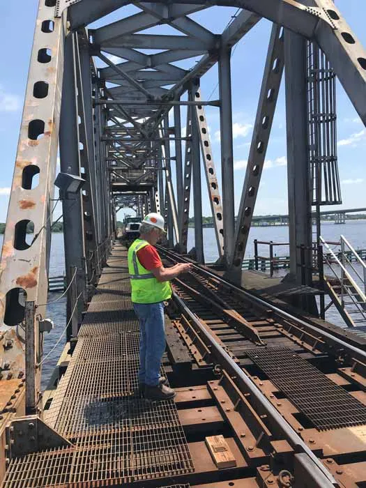 A worker in a hard hat and safety vest on the deck of a steel railroad bridge