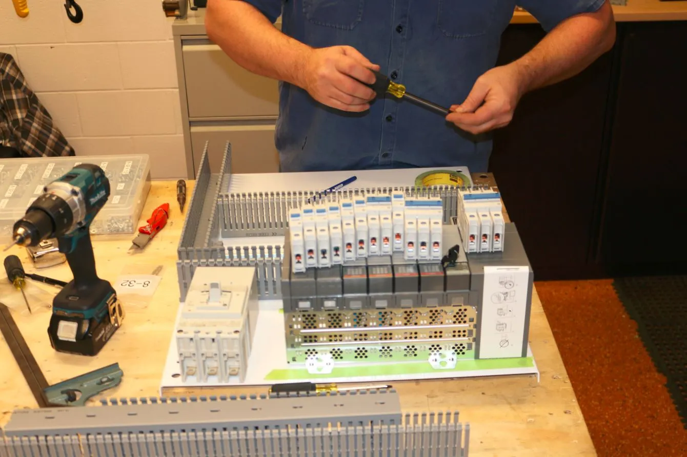 Technician wiring terminal blocks and circuit breakers onto a control panel back plate at a workbench