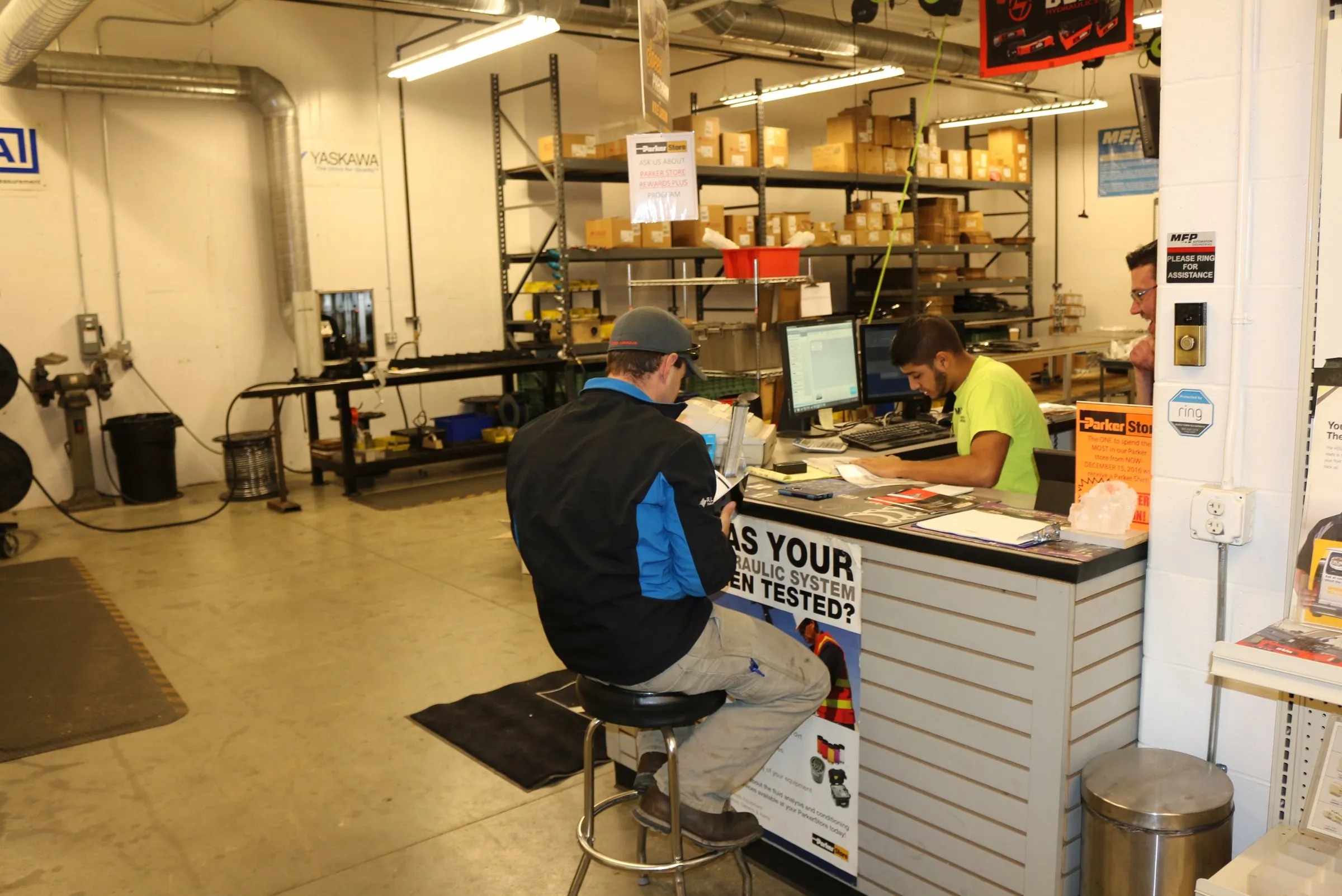 A customer sits at a counter while an MFP employee works at a computer, beside a hydraulic-system testing sign