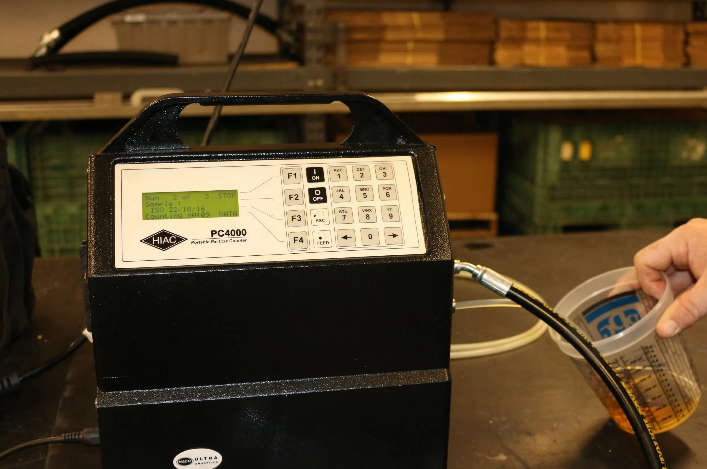 Sample cup of amber oil connected by tube to a HIAC PC4000 particle counter mid-count on its display