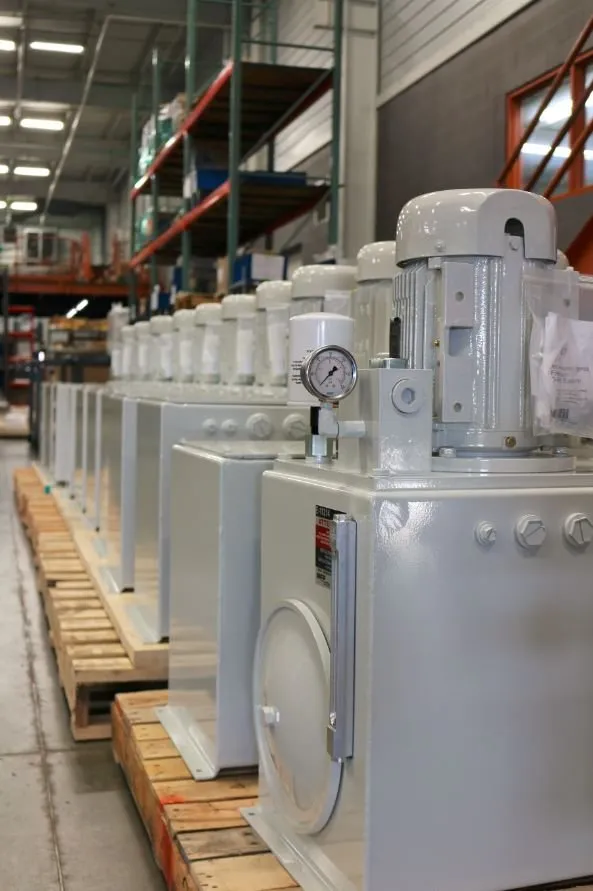 Row of white hydraulic power units on pallets in a warehouse aisle, with pallet racking in the background