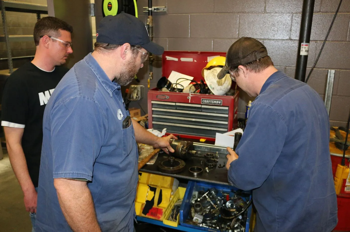 Three men examine a disassembled gear and shaft at a workbench beside a red tool chest and bins of parts, one wearing an MFP shirt