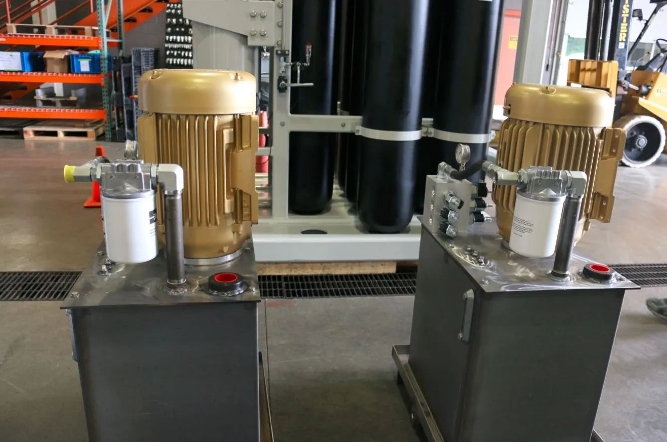 Two stainless steel hydraulic power units with gold motors, in front of black accumulator tanks in a shop bay