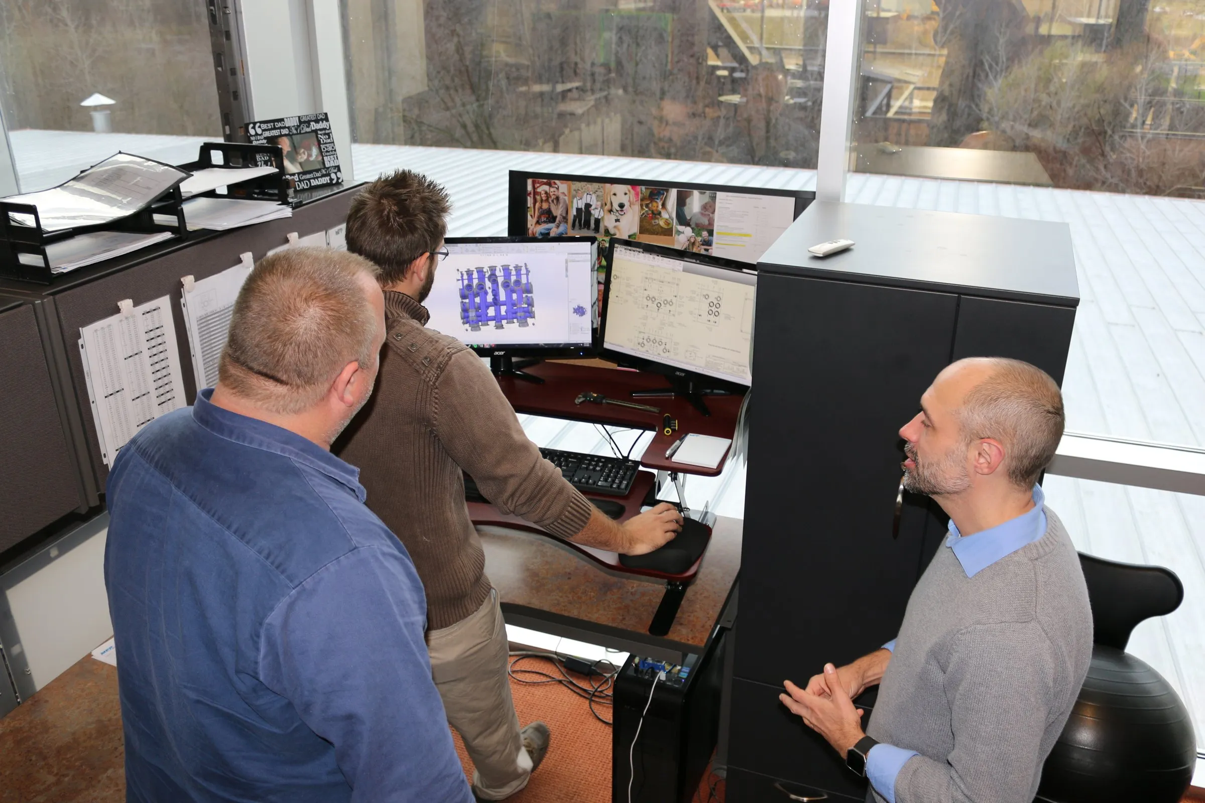 Three men talking beside a CAD workstation in a window-lined engineering office
