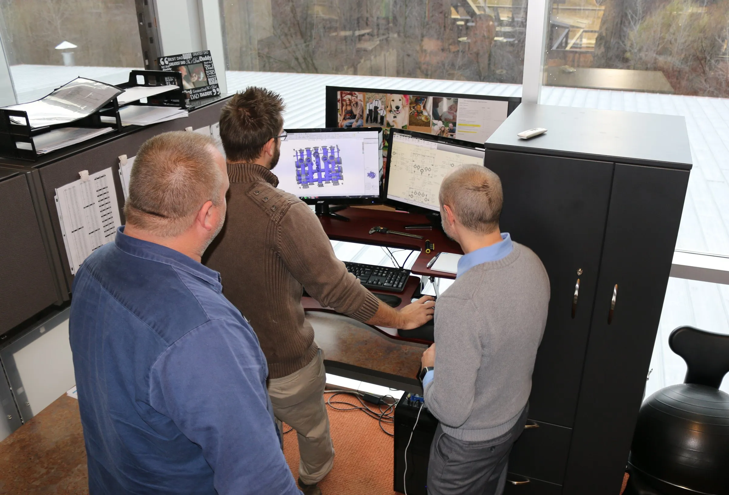 Three people gathered around a workstation reviewing a 3D hydraulic assembly model and drawing