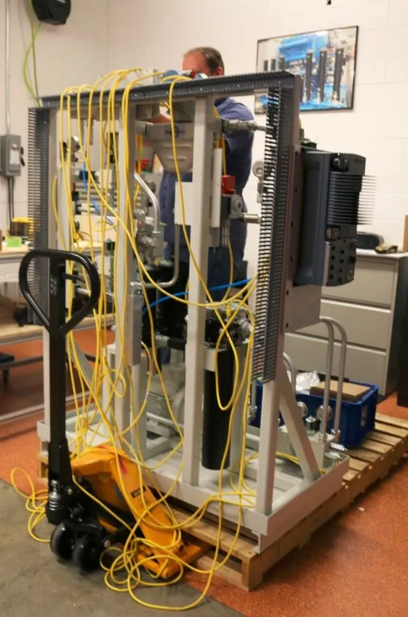 A test skid wrapped in yellow control cables, with a worker checking wiring at the top of the frame