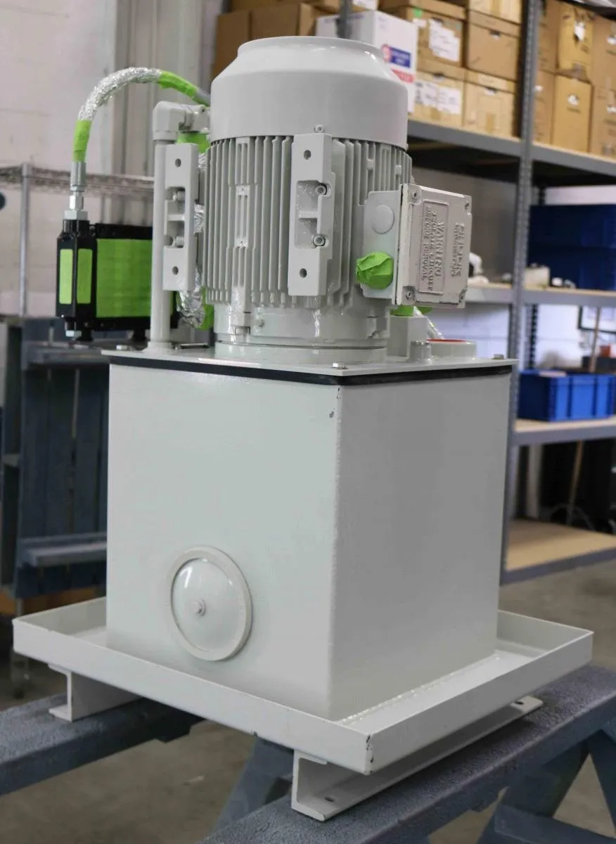 White hydraulic power unit with a motor and green-taped fitting, on a shelf near stacked boxes