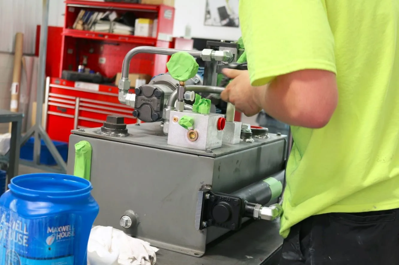 Worker in a safety-green shirt adjusting a fitting on a small hydraulic power unit, with a coffee container nearby
