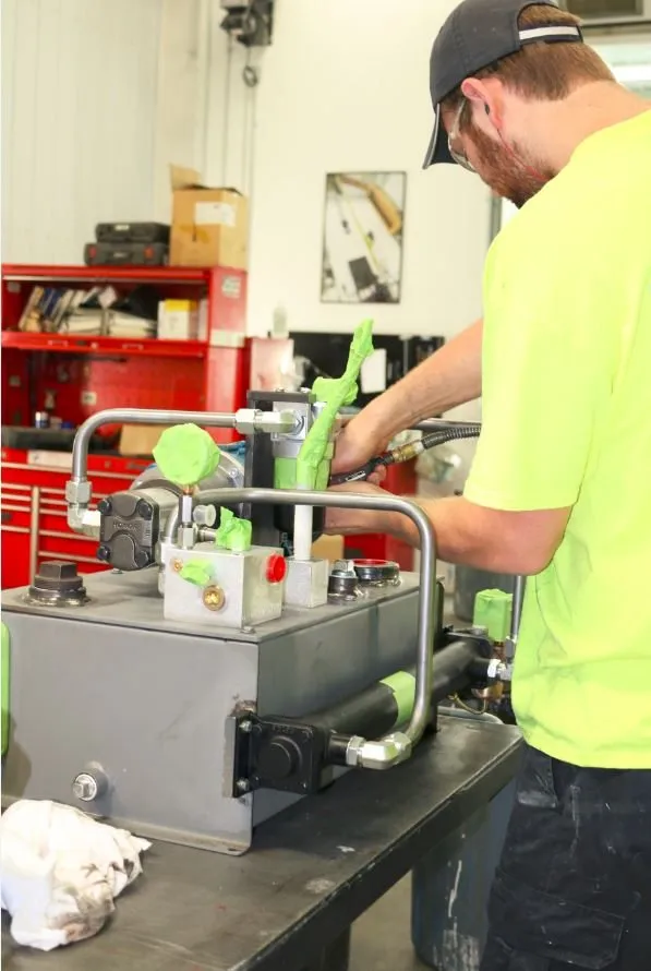 Worker in a cap and safety-green shirt adjusting a fitting on a small hydraulic power unit at a workbench