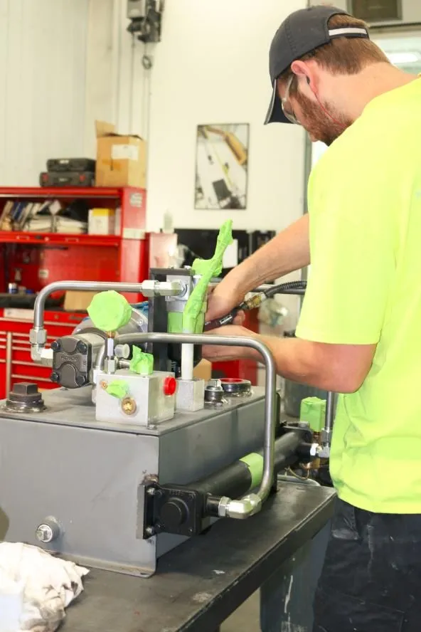 Worker in a cap and safety-green shirt fitting a hose onto a small hydraulic power unit at a workbench