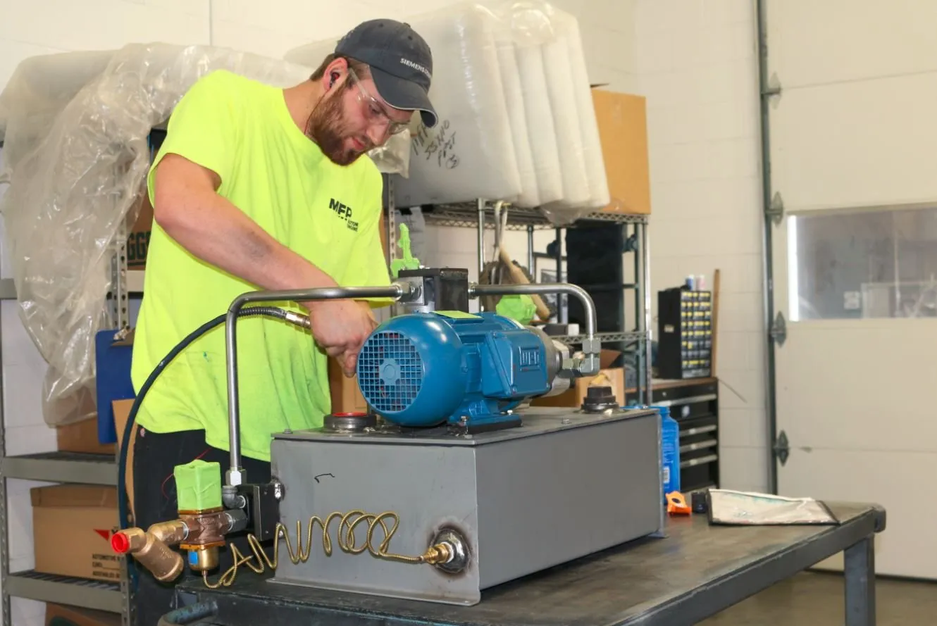 A worker in a yellow MFP safety shirt assembles a blue motor onto a small hydraulic power unit
