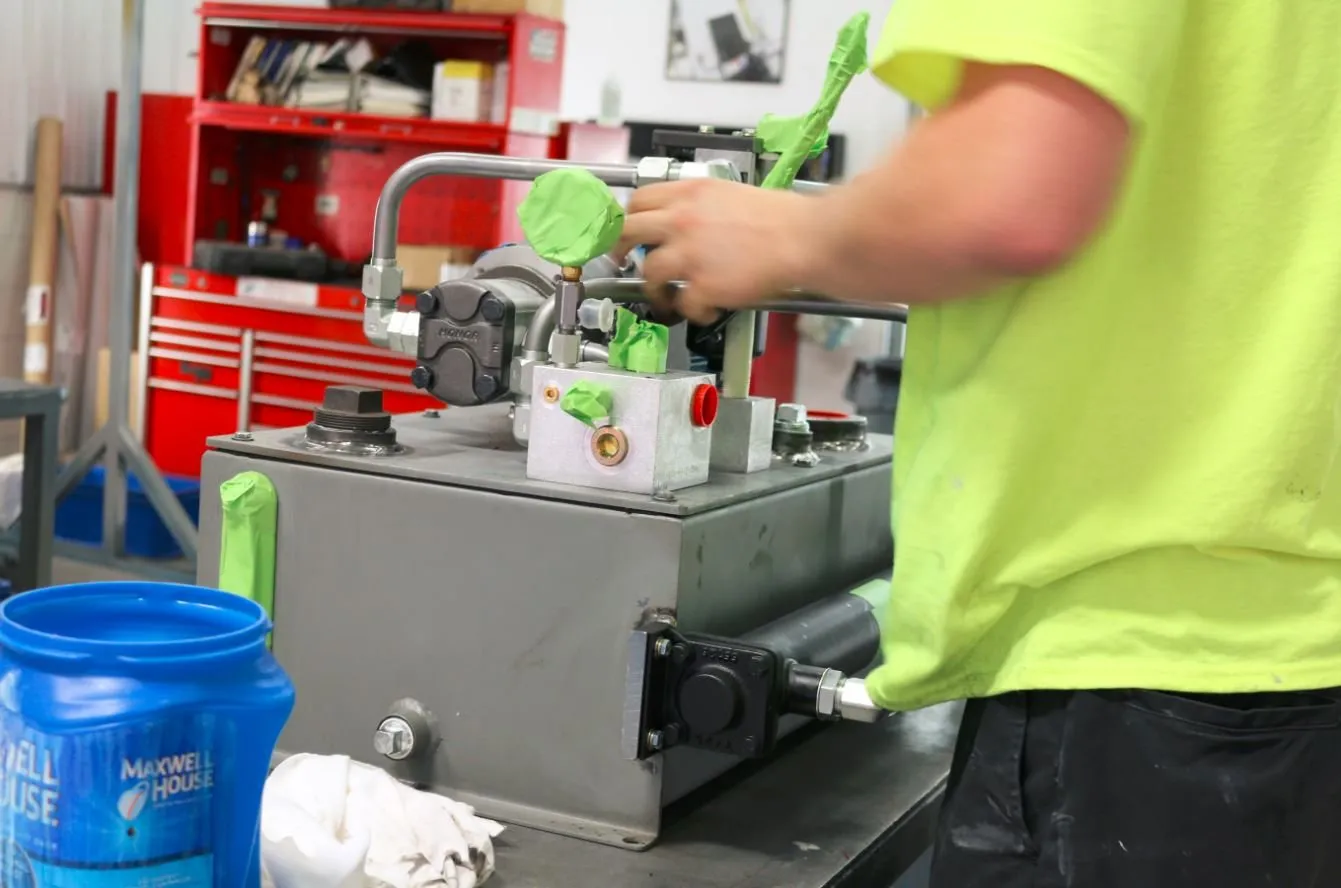 Worker in a safety-green shirt using a wrench on a small hydraulic power unit at a workbench, with a coffee container nearby